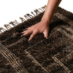 Main sur tapis berbère marron foncé à motifs géométriques en laine épaisse, texture moelleuse et artisanale