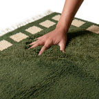 main enfoncée dans tapis berbère vert à carrés beiges, texture épaisse et moelleuse offrant confort premium et douceur